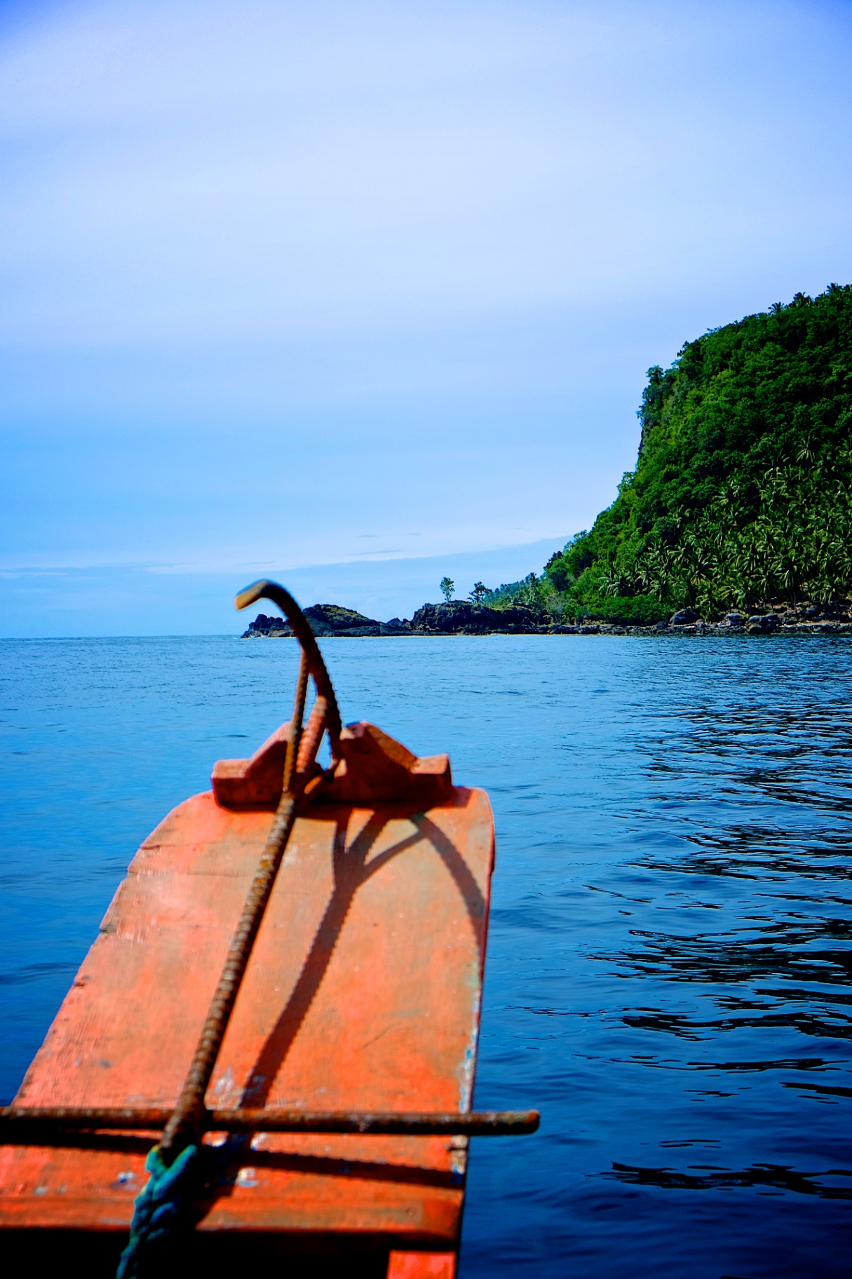 Red bow of a bangka boat cutting toward a lush island shoreline on glassy blue water