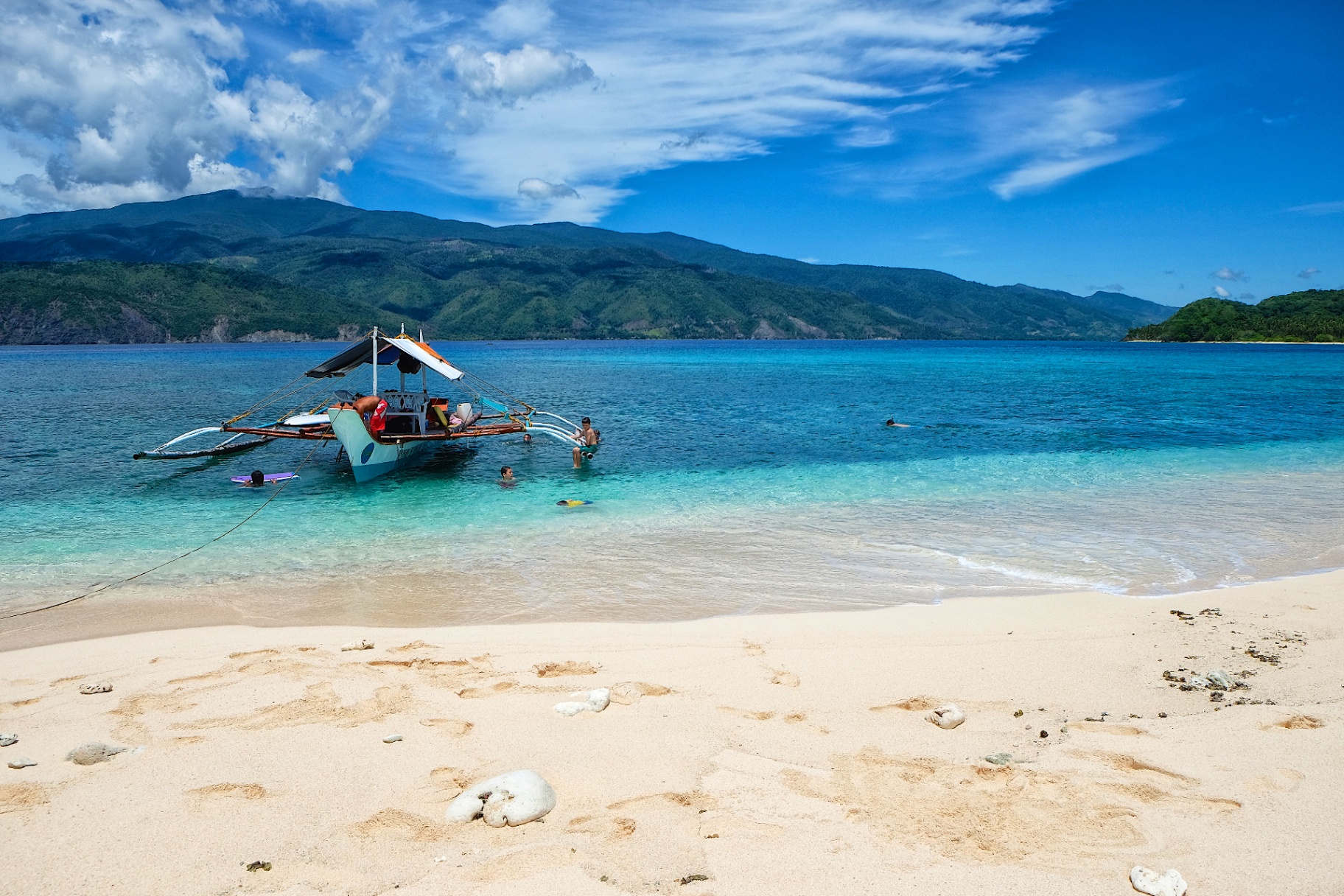 Filipino bangka outrigger anchored in clear turquoise water beside a white sand beach with distant blue mountains