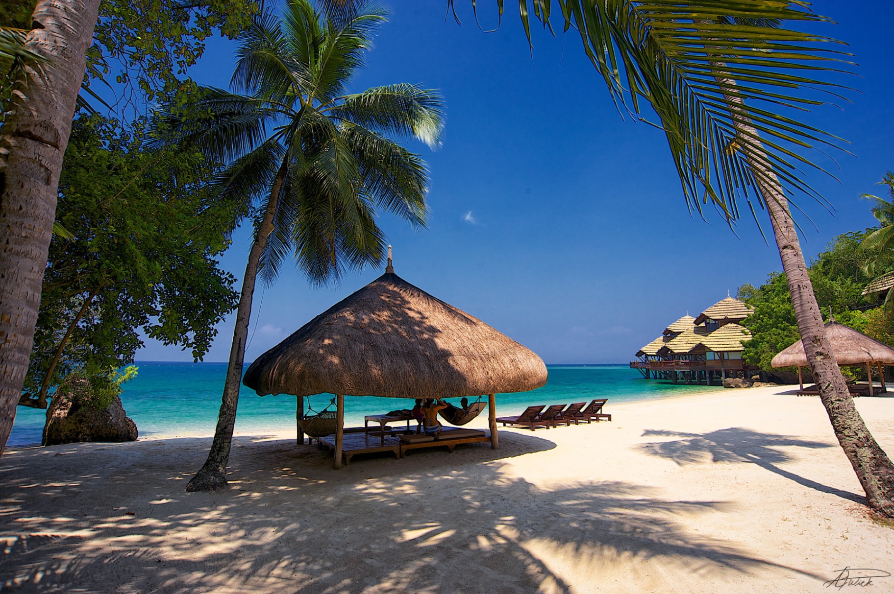 Thatched-roof beach cabana with hammocks framed by coconut palms over white sand and blue Philippine sea