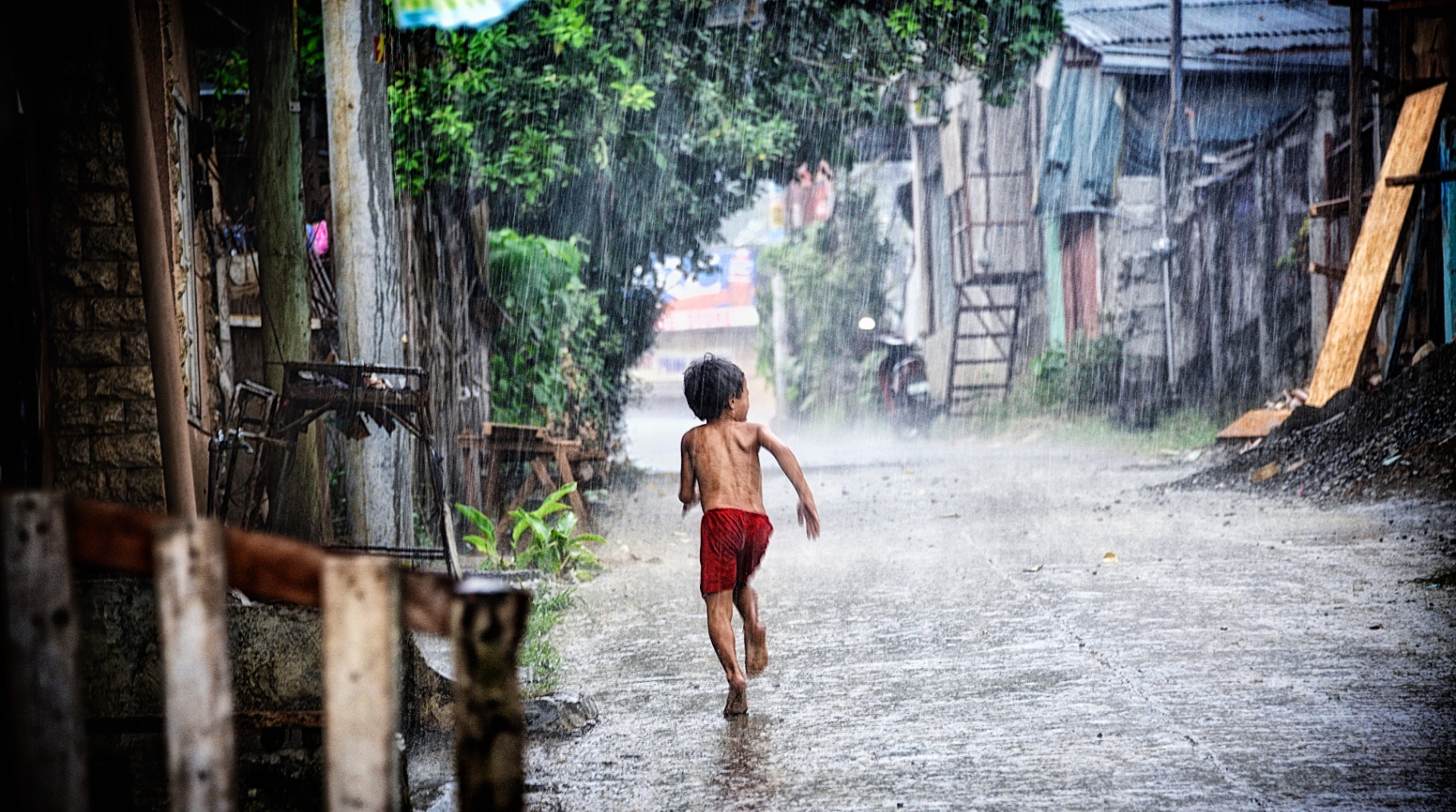 Filipino boy in red shorts running barefoot down a village alley in heavy monsoon rain