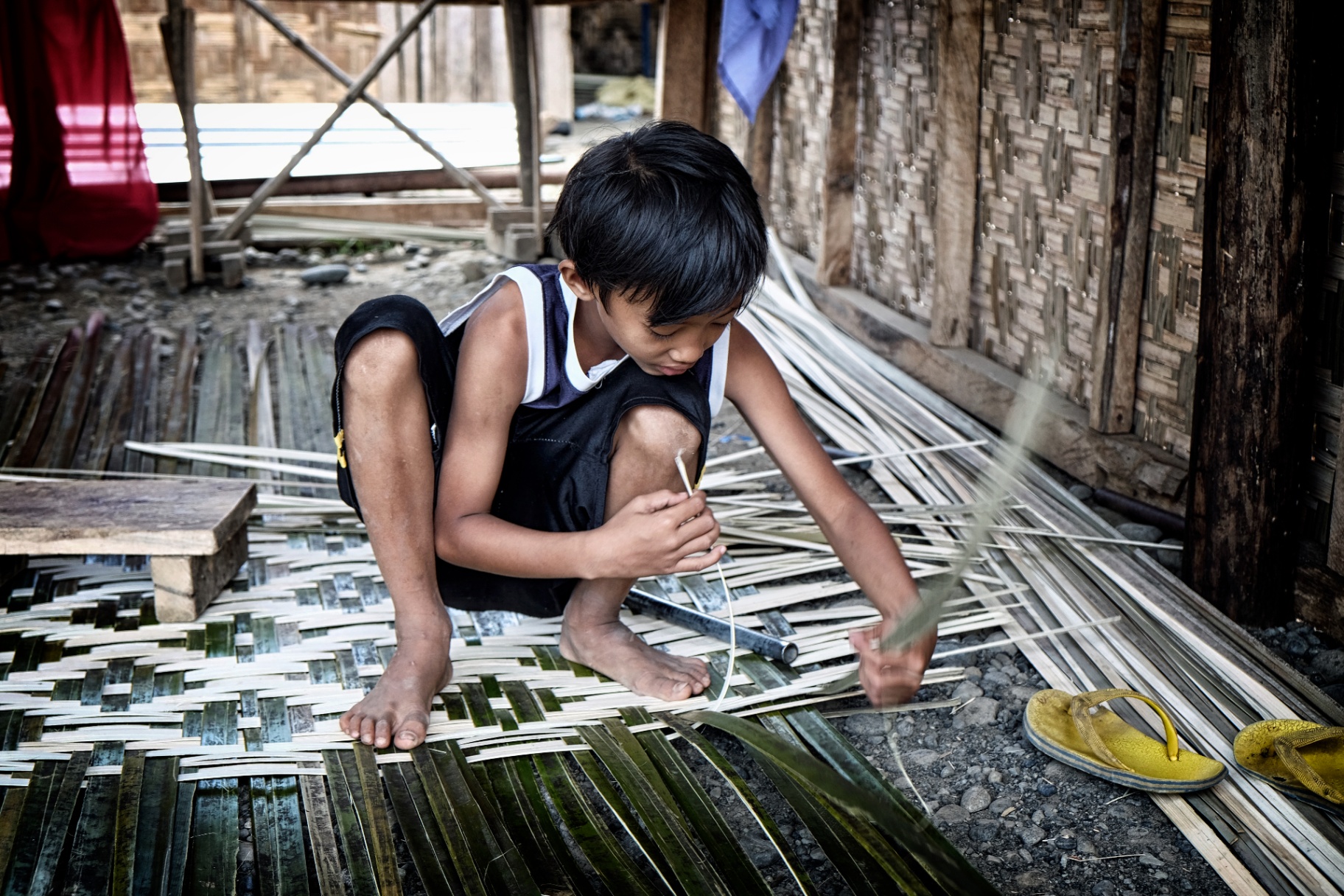 Filipino boy crouched on a bamboo hut floor weaving a palm-leaf mat by hand