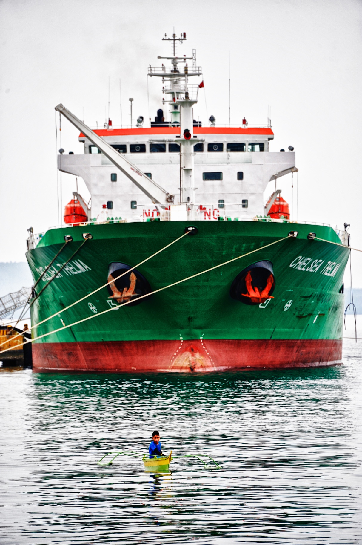 Lone boy in a small bangka outrigger dwarfed by the bow of the cargo ship Chelsea Thelma in a Philippine harbor