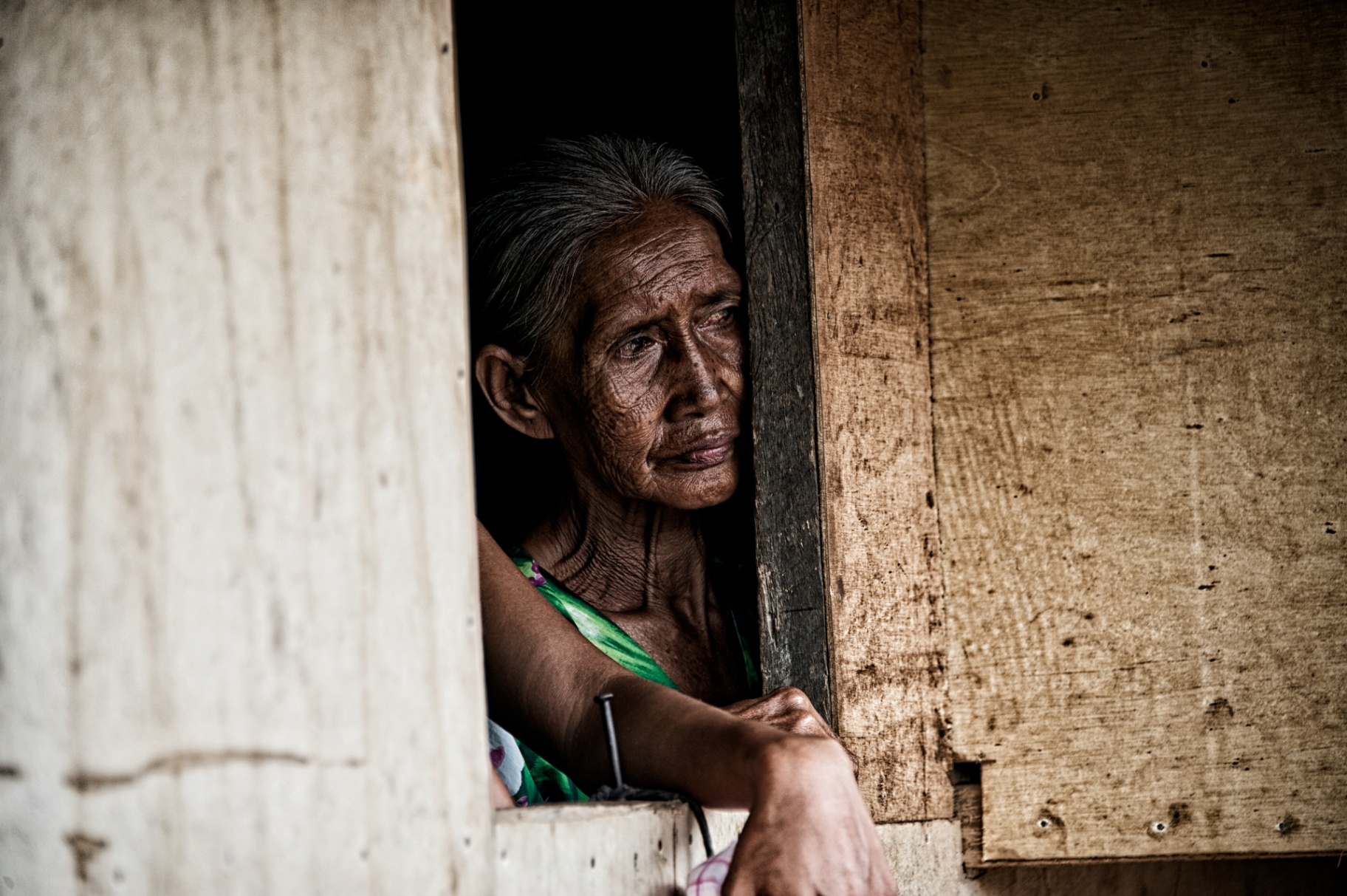 Weathered Filipina elder leaning from the wood-plank window of her small home