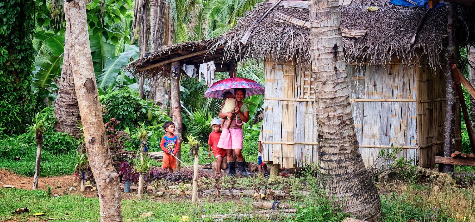 Mother sheltering children beneath a pink umbrella outside a woven nipa home in the rain