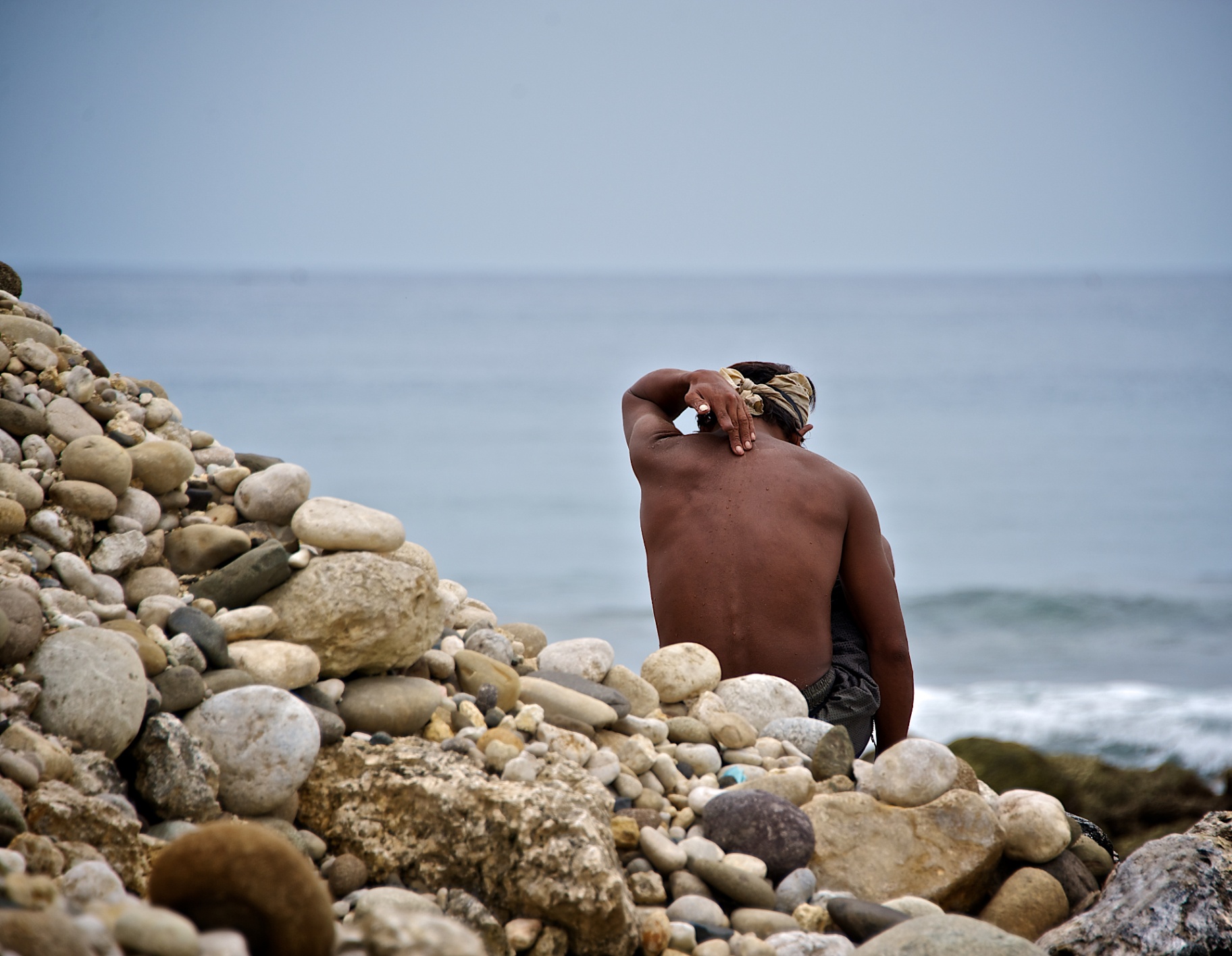 Filipino fisherman with bandana resting on a pile of round shore stones facing the sea
