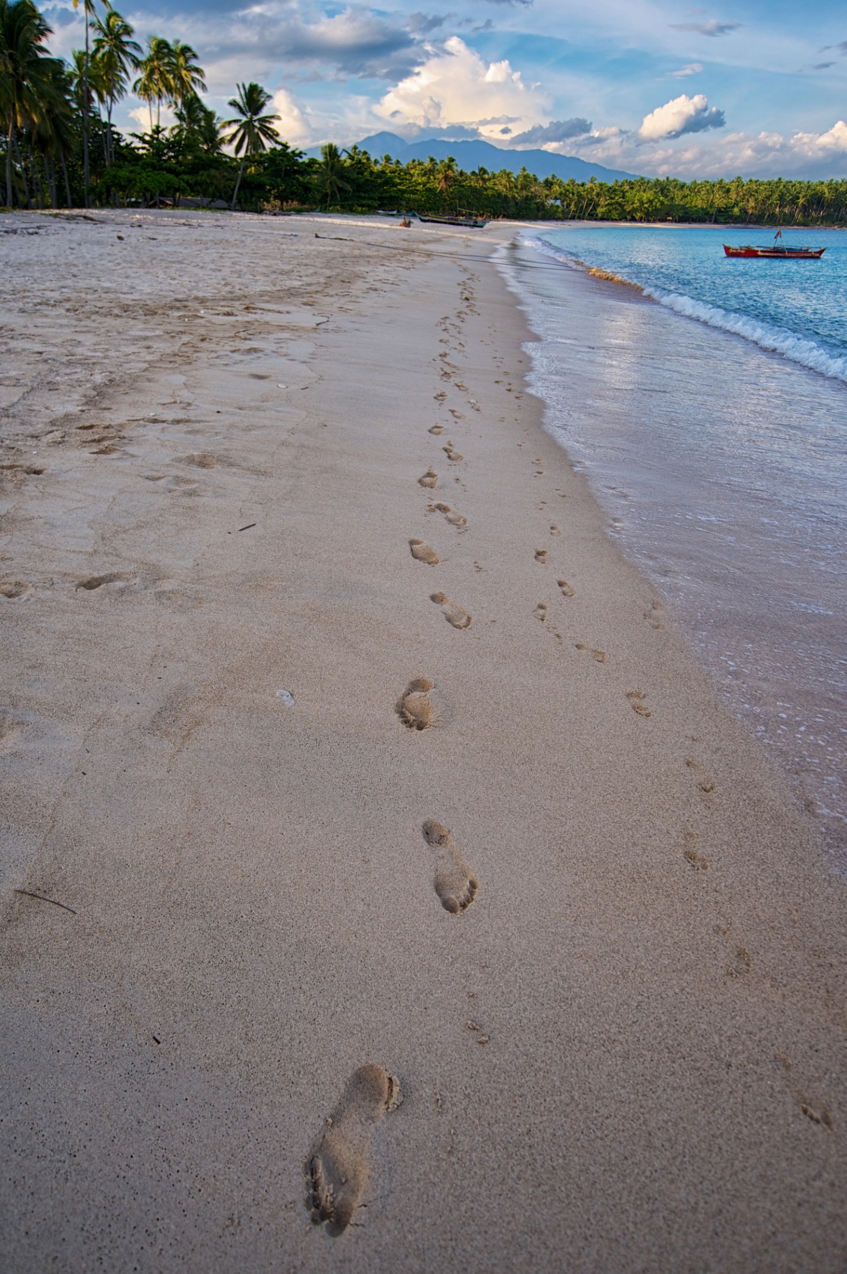 Single trail of footprints along a Philippine shore with a red bangka and distant mountains