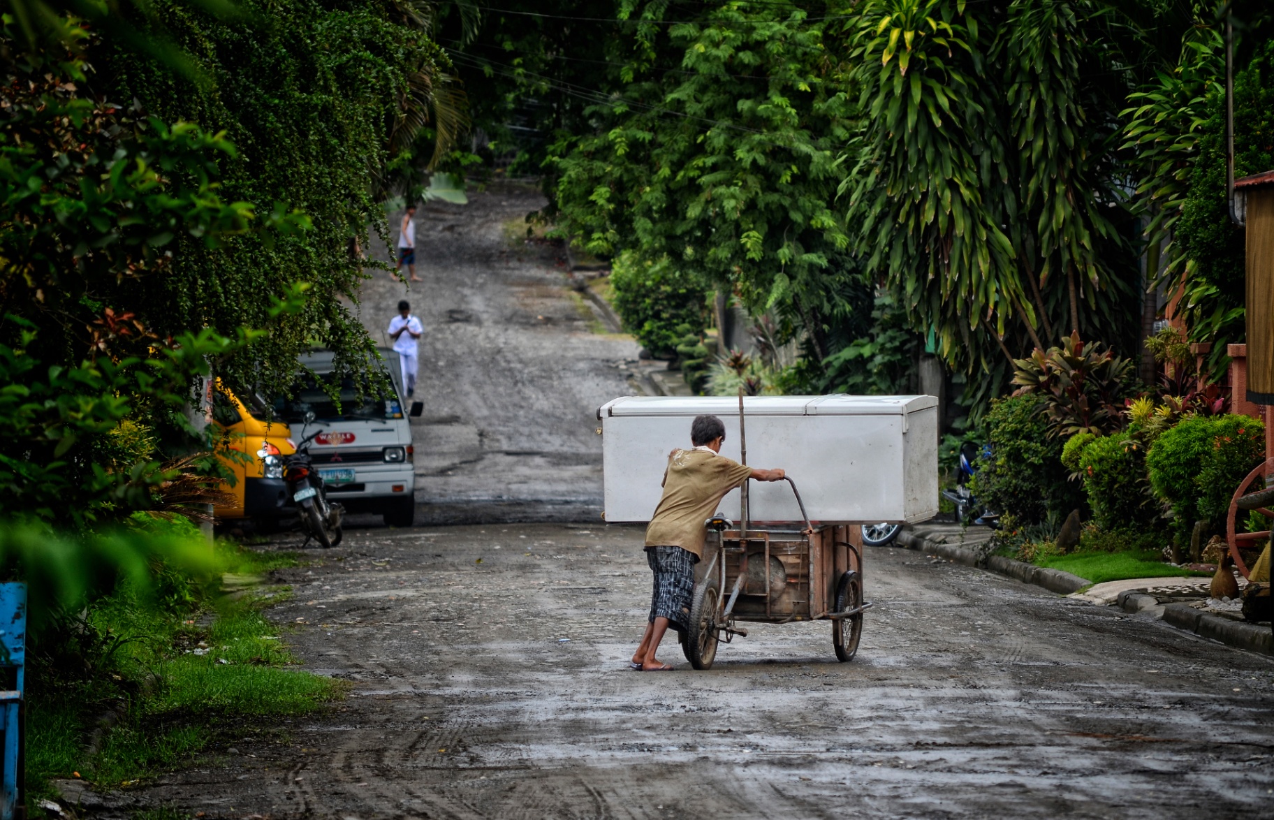Barefoot Filipino boy pushing a wooden tricycle cart carrying a large freezer down a tree-lined village street