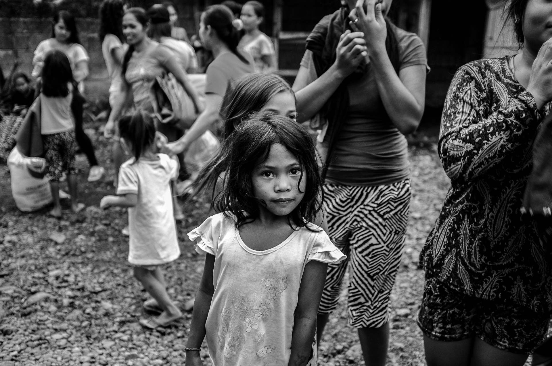 Black and white frame of a young Filipina girl gazing into the camera as a crowd of women and children moves around her