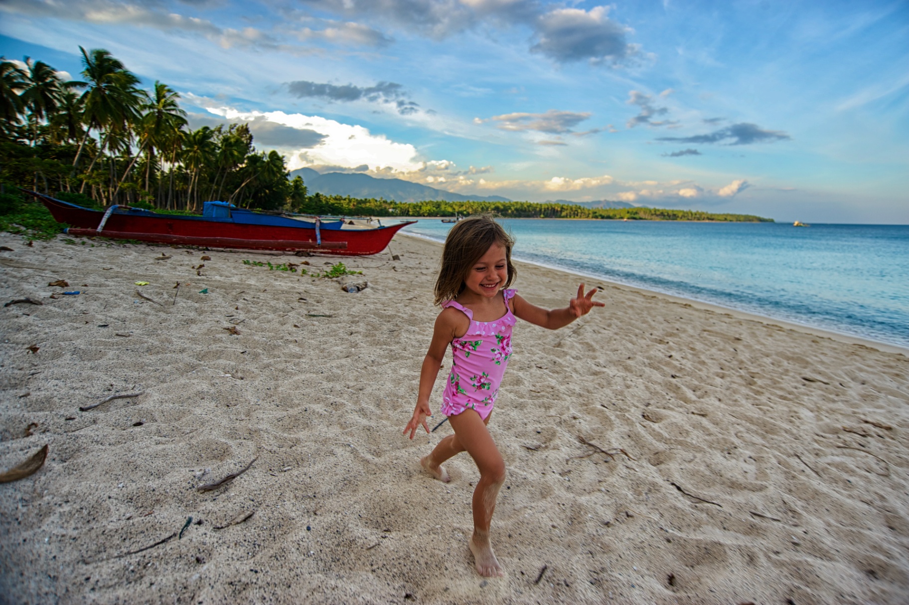 Young girl in a pink swimsuit running barefoot down a Philippine beach past a red bangka boat