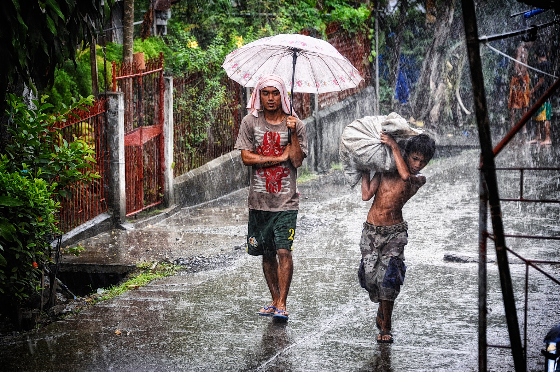 Two Filipino youths walking through monsoon rain, one with an umbrella and one carrying a sack