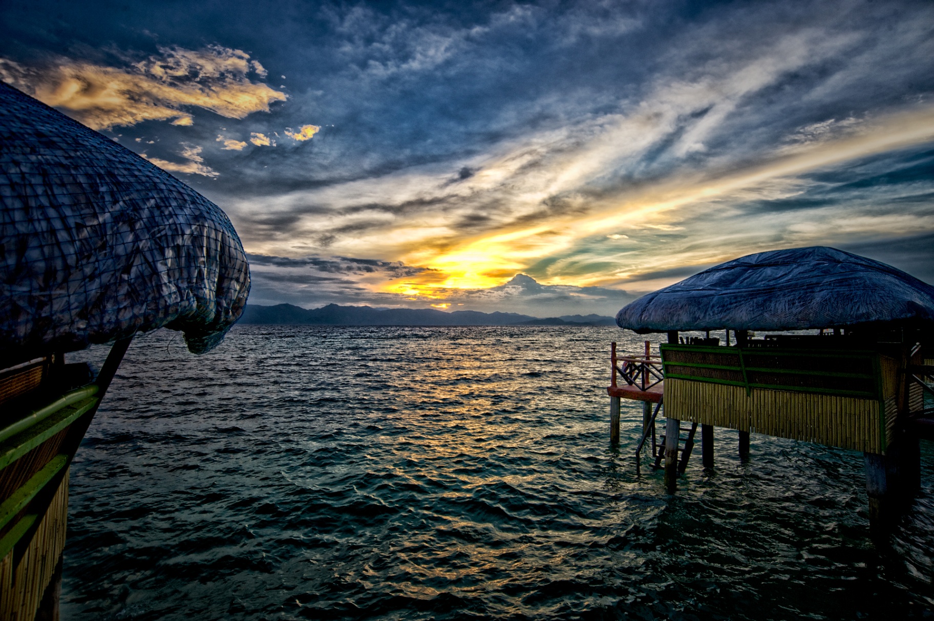 Two overwater nipa huts at sunset on a Philippine sea with distant mountains