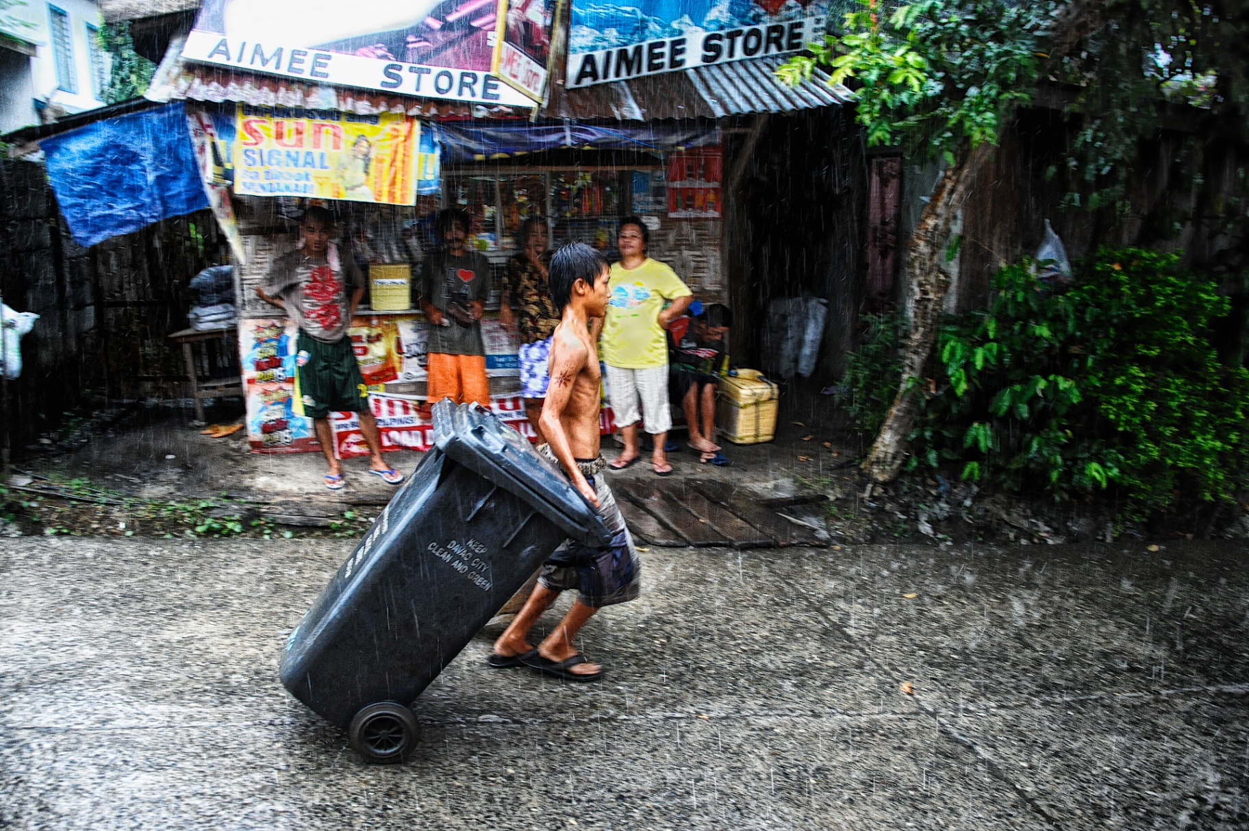 Barefoot Filipino boy hauling a wheeled bin through pouring rain past a roadside Aimee Store in Davao City