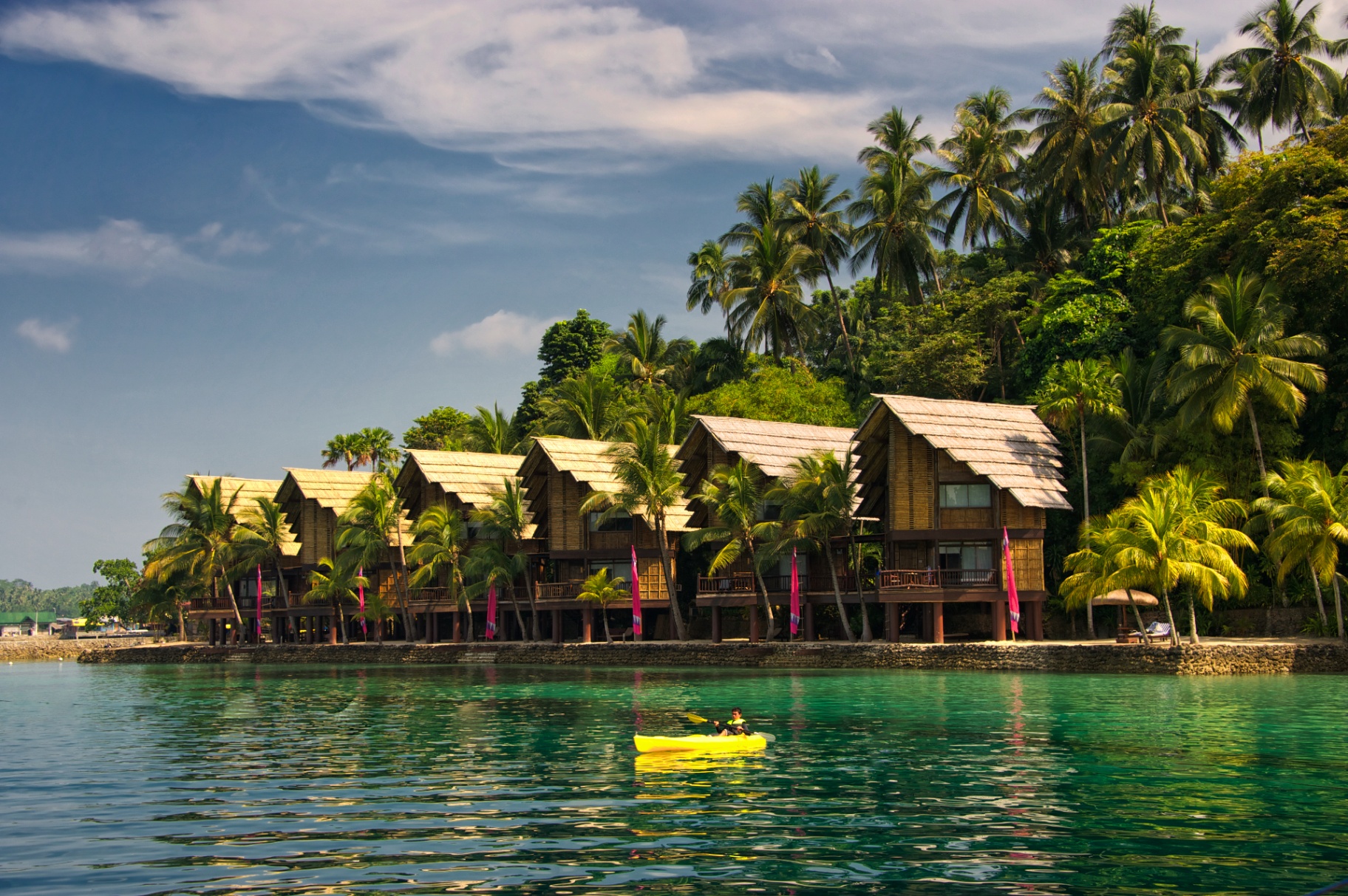 Row of bamboo and thatch bungalows along a palm-lined shore, kayaker on turquoise water in front