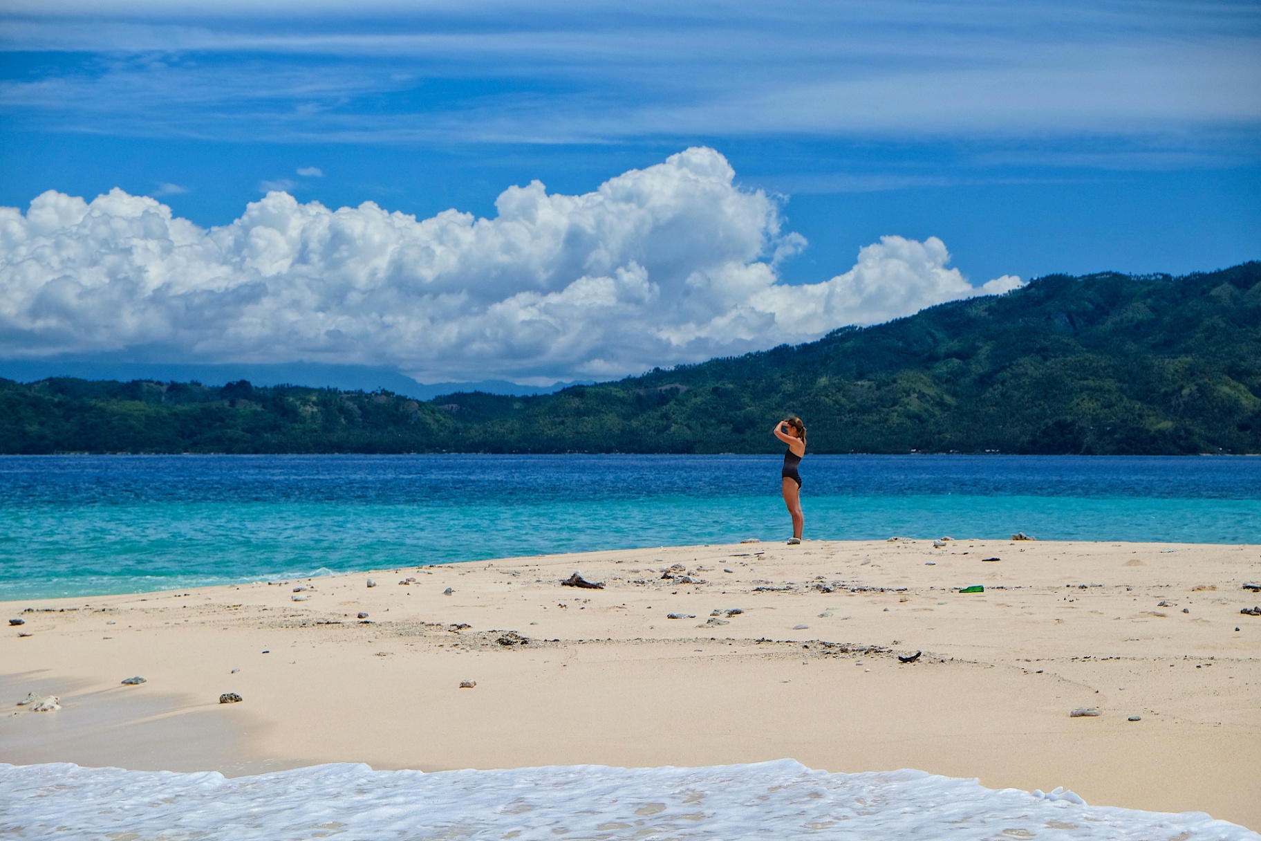 Solitary woman standing on a luminous sandbar with turquoise shallows and distant green mountains