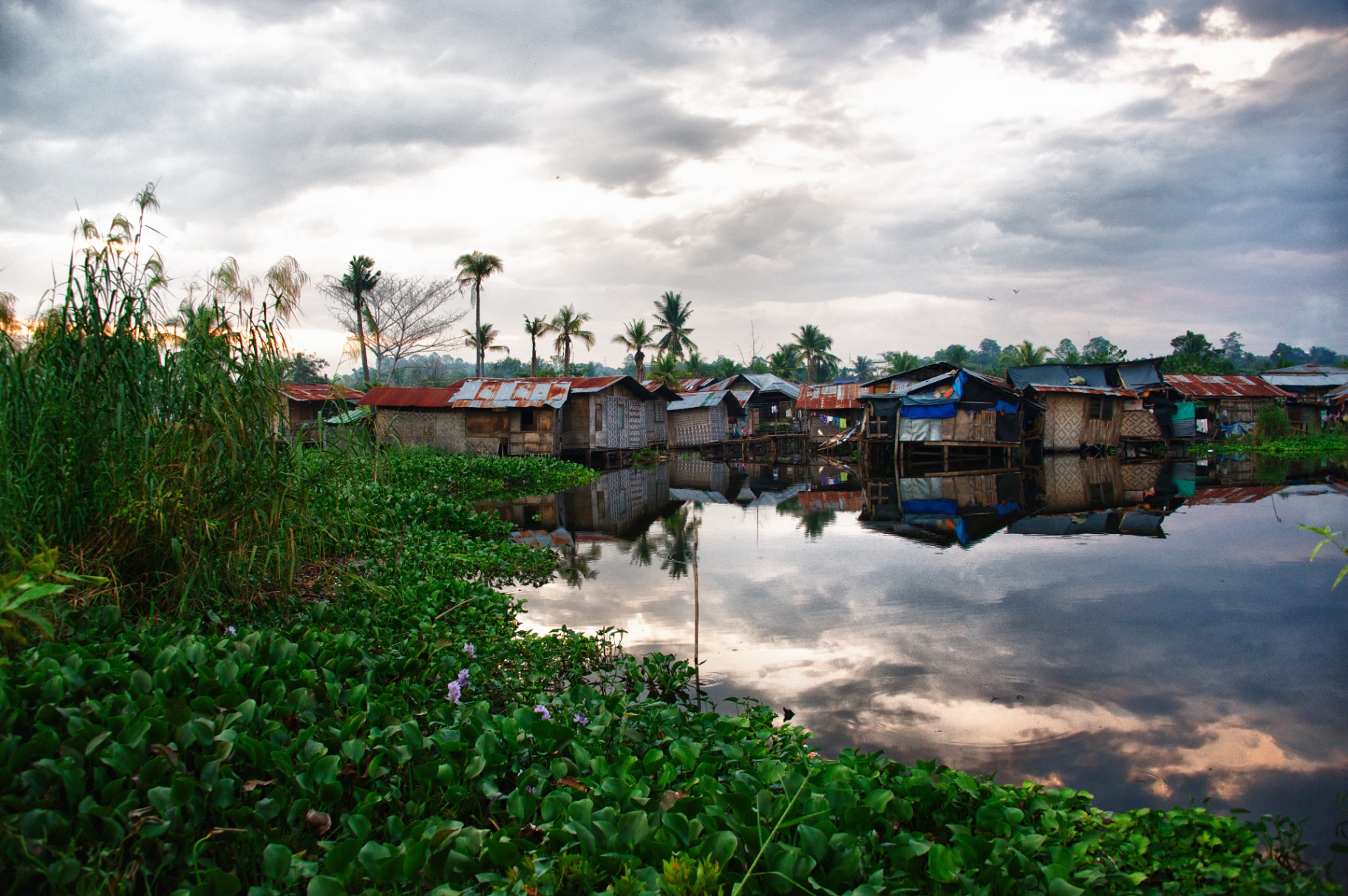 Stilt-house fishing village mirrored in still water at dawn beneath a dramatic cloud-streaked Philippine sky