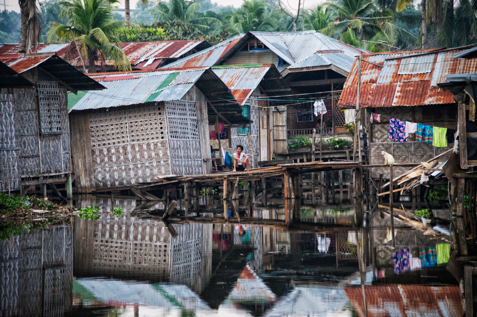Woman seated along a timber walkway in a stilt-house village reflected in still water