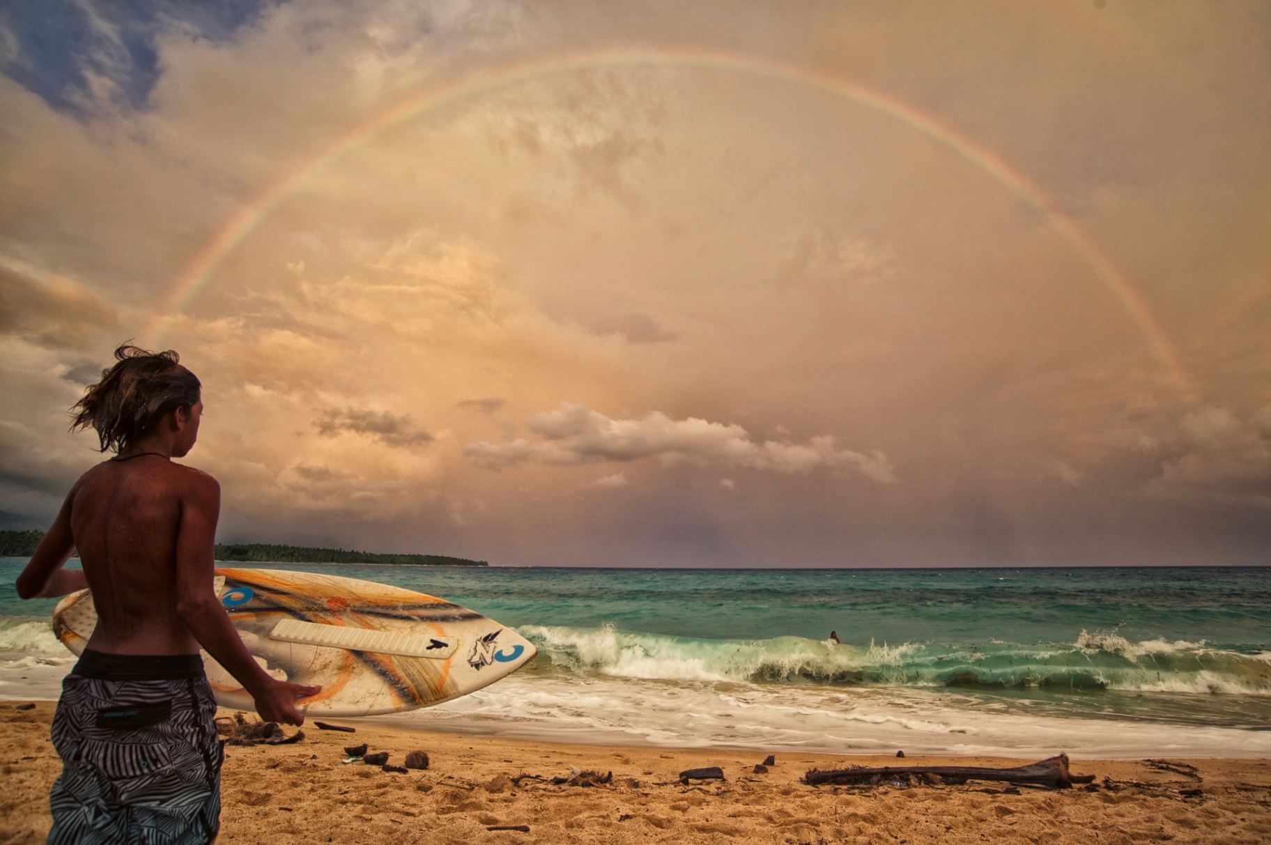 Young surfer carrying a board toward the surf under a full rainbow, Philippines